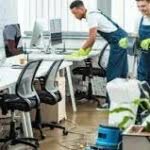 3 uomini addetti alle pulizie in divisa salopette blu e maglietta bianca puliscono un ufficiocomposto da una lunga scrivania con tante sedie
